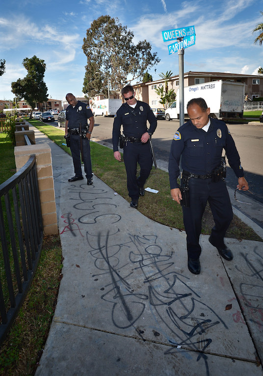 Huntington Beach Police Officers Bernard Atkins, left, Sean McDonough and Juan Munoz look over gang graffiti on the sidewalk at the corner of Queens Lane and and Barton Drive in Huntington Beach that had been crossed off and replaced with new gang related graffiti. The officers are part of a new effort to build community relationships and reduce gang violence in the Oak View neighborhood of Huntington Beach. Photo by Steven Georges/Behind the Badge OC