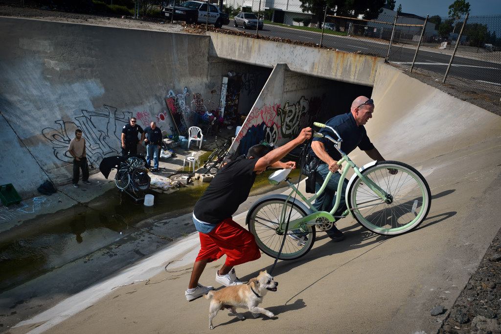 A Garden Grove PD officer helps a homeless man and his dog living in a flood canal under Western Ave. in Garden Grove up the steep embankment as the Garden Grove Homeless Task Force conducts a cleanup sweep. Photo by Steven Georges/Behind the Badge OC