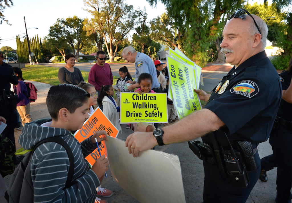 As kids arrive at Pepper Tree Park in Tustin for the morning walk, Capt. Steve Lewis hands out safety posters for select kids to hold up during Tustin PD’s annual Walk to School campaign. Photo by Steven Georges/Behind the Badge OC