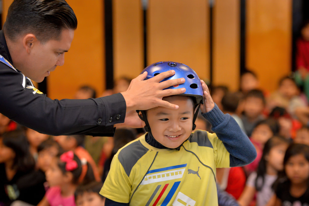 Garden Grove PD Officer Royce Wimmer helps Briman Tran, 6, demonstrate the proper way to put on a bicycle helmet during a safety assembly at Anderson Elementary School in Garden Grove. Photo by Steven Georges/Behind the Badge OC
