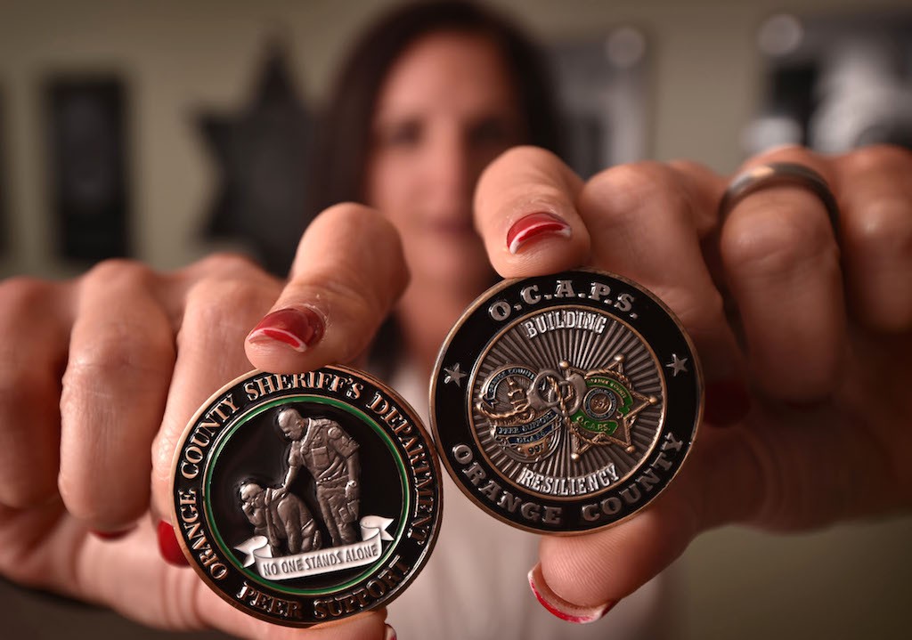 Heather Williams, Regional Peer Support Coordinator for the Orange County Sheriff’s Department, with the Orange County Sheriff’s Challenge Coin, left, for Pier Support with the theme No One Stands Alone. On the right is the Orange County Association Of Peer Supporters or OCAPS coin. The graphic on the right represents, according to Williams, “Law Enforcement of Orange County, intertwined and working together in the event that a department needs mutual aid or support in the aftermath of a critical incident.” Photo by Steven Georges/Behind the Badge OC