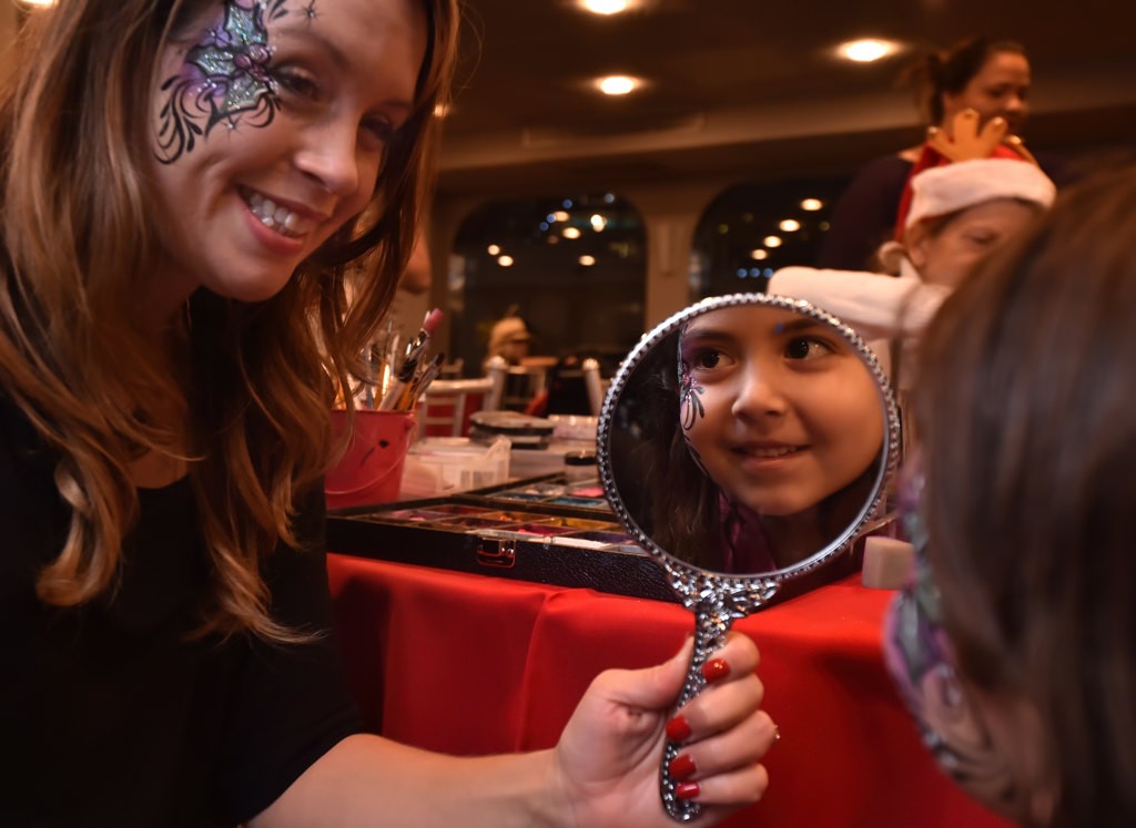 Face painter Sara Sanchez holds a mirror up to Savannah Hernandez, 5, to view her facial artwork during the Make A Wish Holiday Harbor Cruise. Photo by Steven Georges/Behind the Badge OC